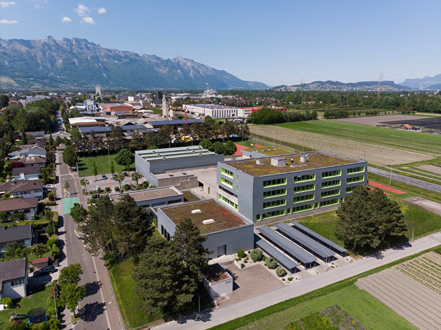 Luftbild der Schulanlage Oberstufenzentrum Flös in Buchs SG, Schulgebäude mit begrüntem Flachdach zwischen Wohnquartier und Feldern, dahinter das Rheintal und die Berge
